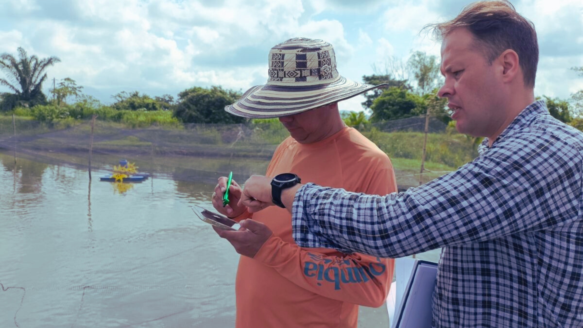 An FAI consultant shows a farmer how to use the Tilapia Welfare App beside a tilapia pond. The consultant points at the app's dashboard displayed on a smartphone held by the farmer.